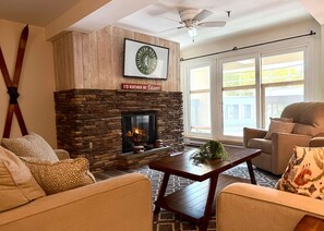A cozy living room with a stone fireplace, a ceiling fan, and a large window with a view of the outside. - BearsLair- Pool, Hot tub, Bike to Mountain! (Killington)