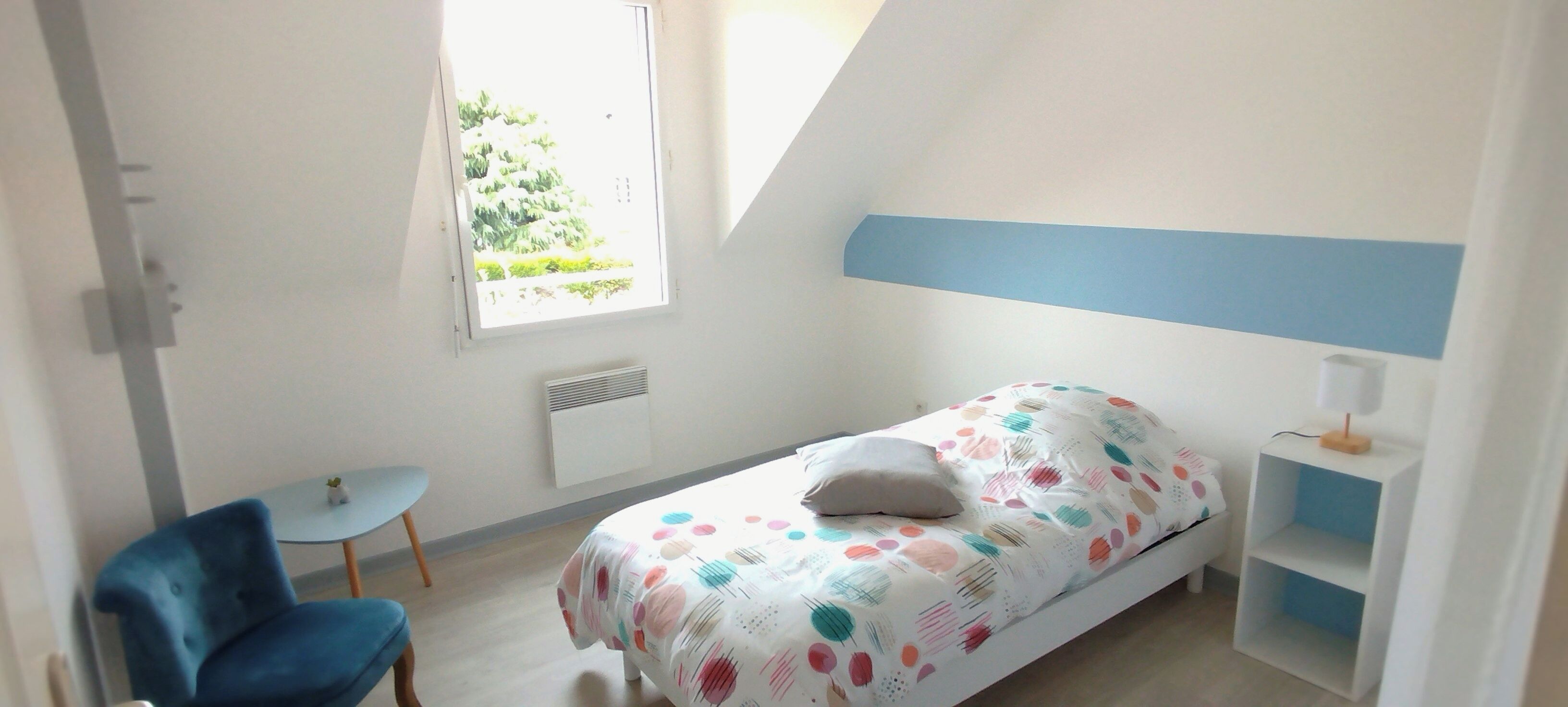 A bedroom with a bed, a chair, a small table, a window, and a shelf.