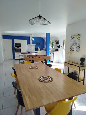 A modern kitchen with white cabinets, a blue accent wall, and a dining area with a wooden table and yellow chairs. - Cap Rance With family and friends! (Plouër-sur-Rance)