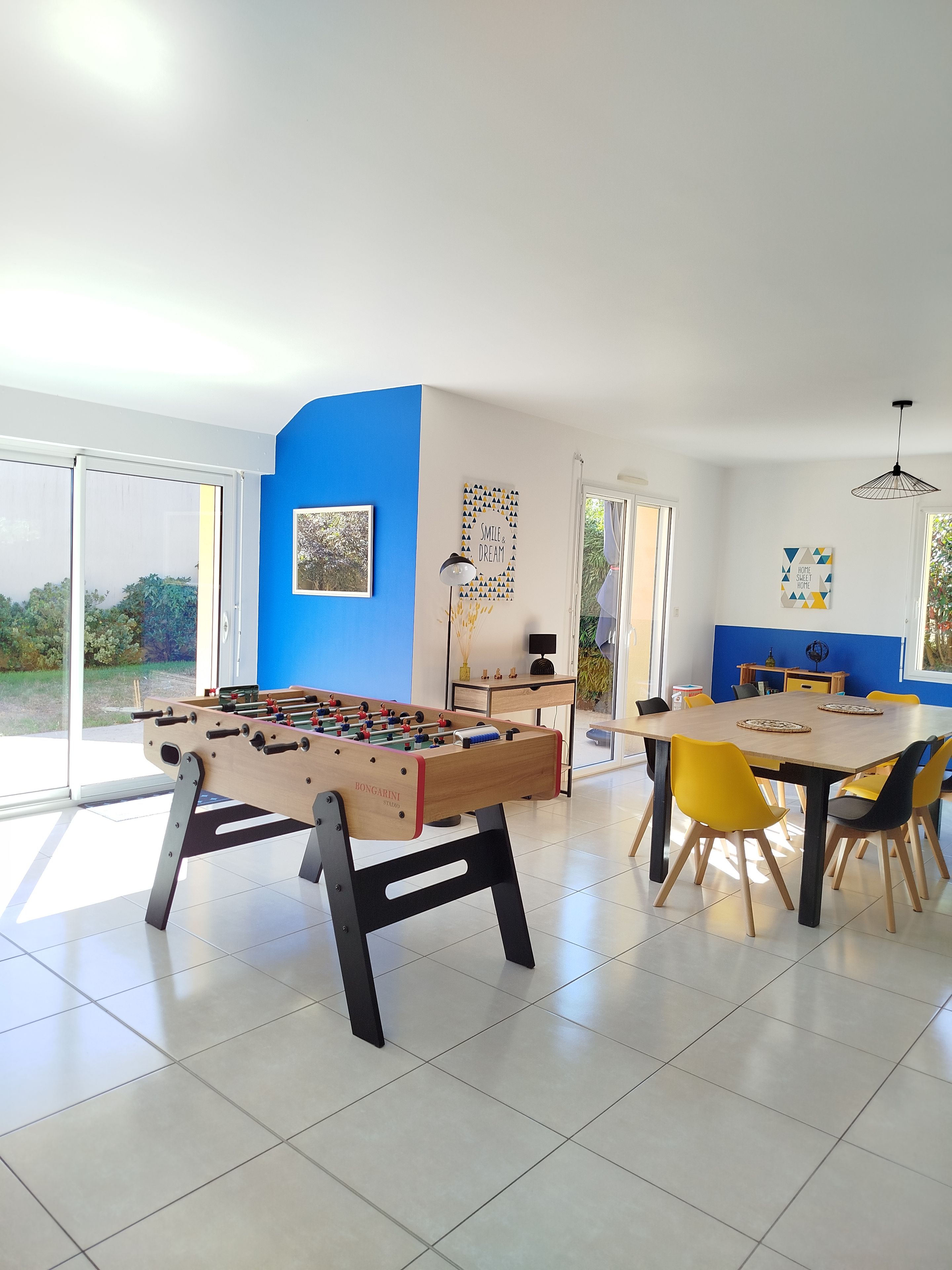 A modern room with a foosball table, a dining table with chairs, and a view through large glass doors.