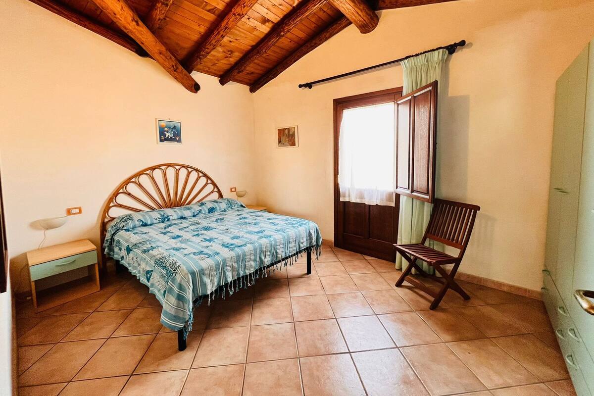 Ein Schlafzimmer mit Holzbalkendecke, einem Bett mit blauer Musterdecke, einem Holzstuhl, einem Nachttisch und einem Fenster mit Vorhang.