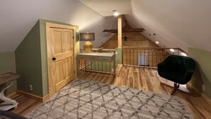 A room with a sloped ceiling, a wooden door, a small table with a lamp, a green chair, and a patterned rug. - Relax & Recharge at Firefly Cabin (Russellville)
