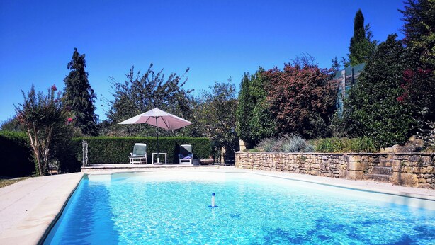 Ein klares, blaues Schwimmbecken mit Liegestühlen und einem Sonnenschirm, umgeben von Steinmauern und viel Grün.