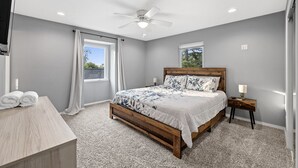 A bedroom with a wooden bed, a ceiling fan, a window with curtains, a bedside table with a lamp, and a small wooden dresser. - Lake Retreat w/ Hot Tub, Game Room & Grill (Rogers)