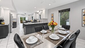 A modern kitchen with dark cabinetry, a central island, and a dining area with a table set for four. - Lake Retreat w/ Hot Tub, Game Room & Grill (Rogers)