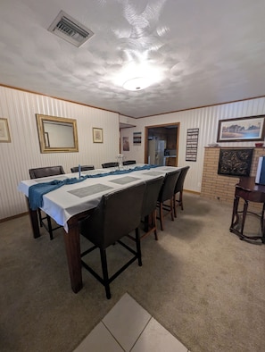 Comedor con una larga mesa, sillas, ventilador de techo y una cocina visible a través de un vano abierto.