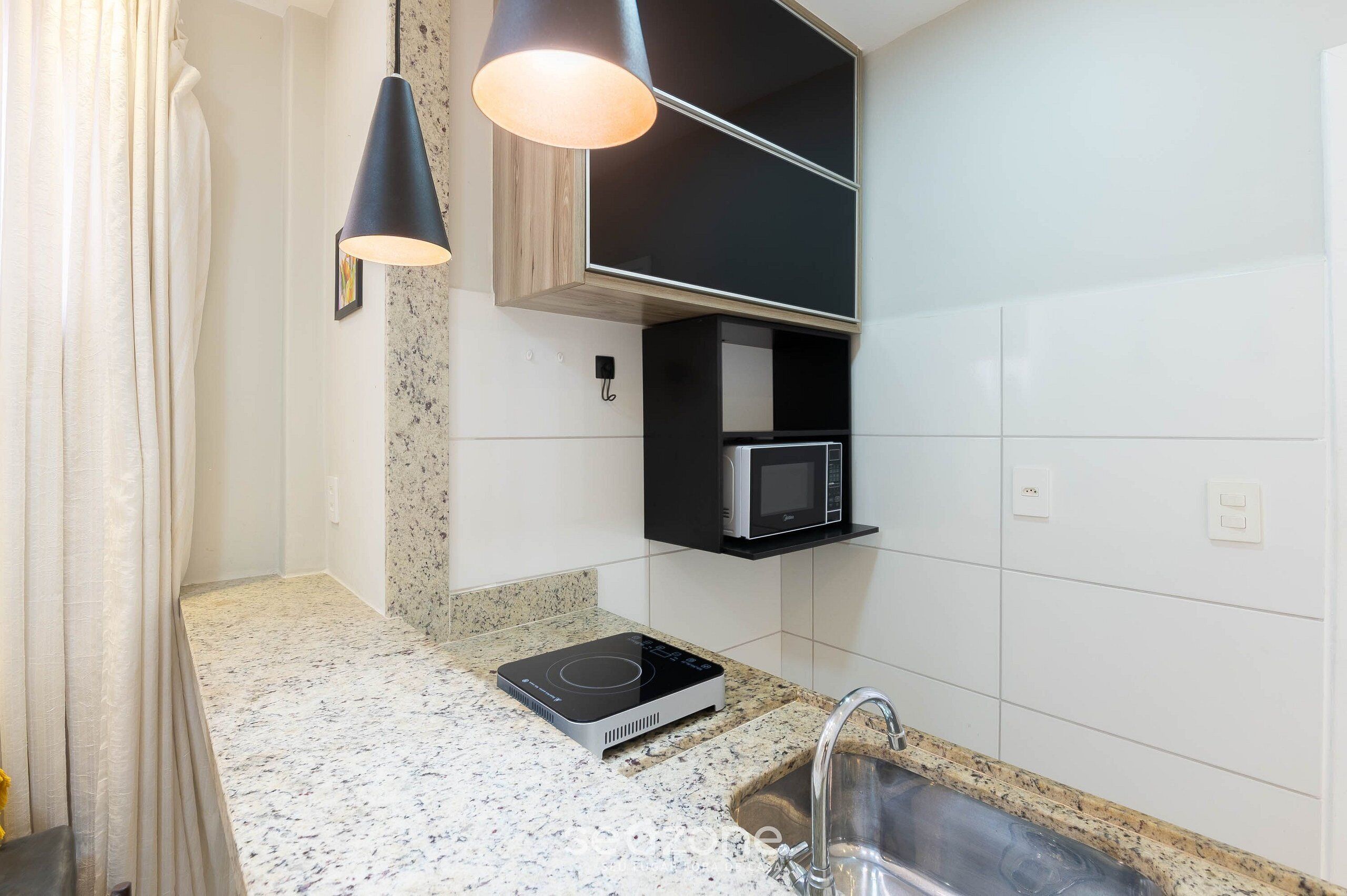 A kitchen with a granite countertop, a built-in microwave, a black hanging light fixture, and a stainless steel sink.