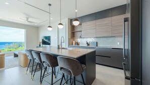 A modern kitchen with a large island, bar stools, and a view of the ocean. - Point West Premier Rentals (West Bay)