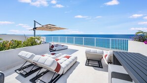 A rooftop terrace with sun loungers, a table, and a large umbrella. The terrace overlooks a clear blue sky and a distant coastline. - Point West Premier Rentals (West Bay)