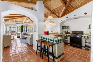Una cocina amplia con una isla central, un comedor con sofá y sillas, y vistas a través de un arco abierto.
