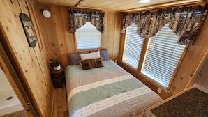 Un dormitorio de cabaña de madera acogedor, con una cama, una mesita de noche, una ventana con persianas y un cojín decorativo.