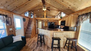 Interior de una cabaña de madera con cocina, una escalera y una sala de estar con sofá y taburetes.
