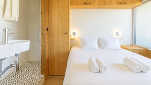 Ein Hotelzimmer mit Holzschrank, einem weißen Bett mit gerollten Handtüchern, einem Badezimmer mit Waschbecken und Dusche sowie einem Fenster mit Tageslicht.