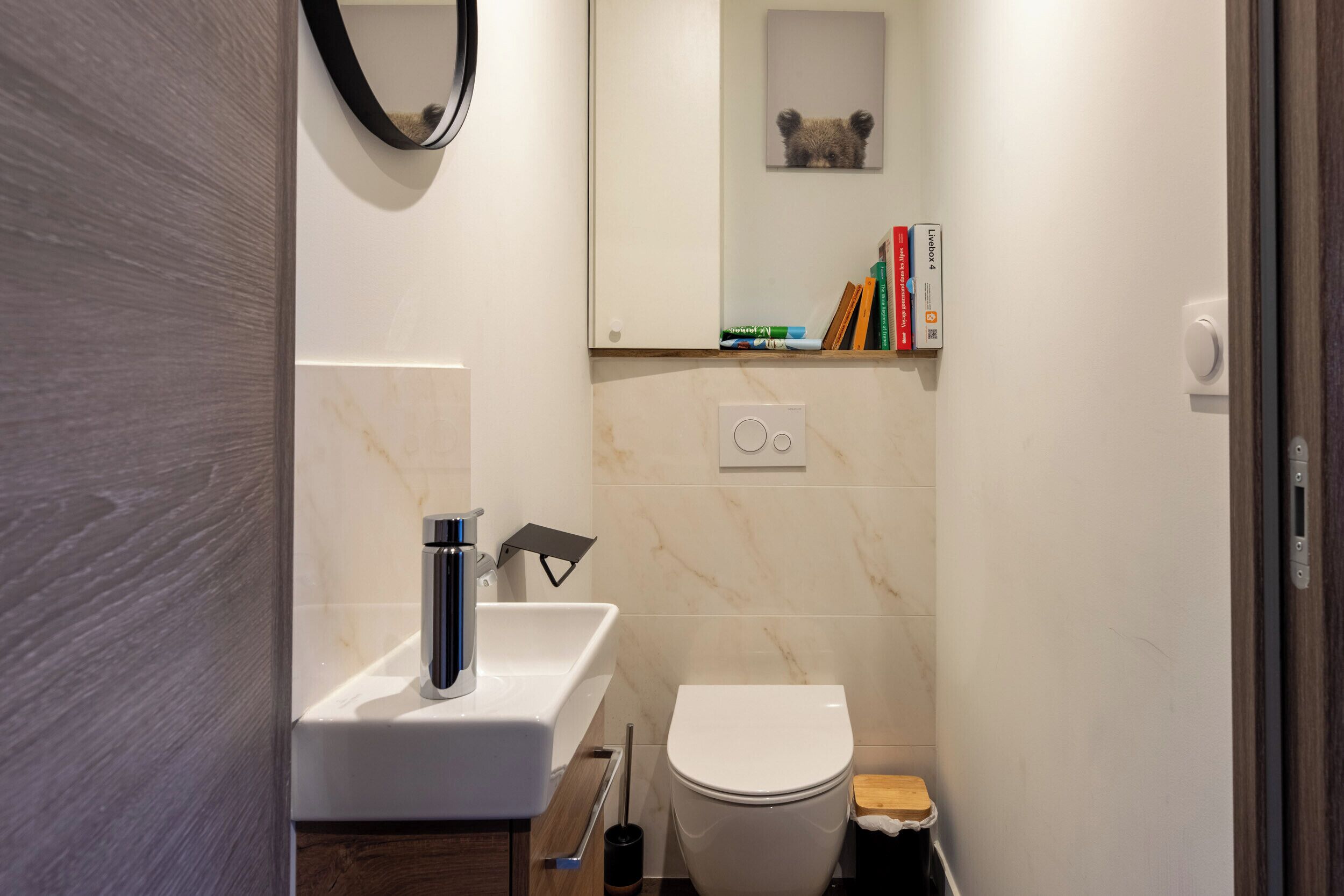 Une petite salle de bain avec un lavabo blanc, des toilettes et une petite étagère contenant des livres et des articles de toilette.