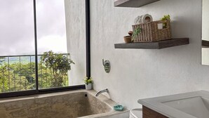 A bathroom with a large window, a bathtub, a toilet with a floral design, and shelves with plants and toiletries. - Eco Suite 1-bedroom villa in awesome La Fortuna with AC (La Fortuna)