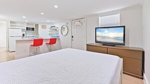 A bedroom with a large bed, a television on a dresser, and a kitchenette in the background.