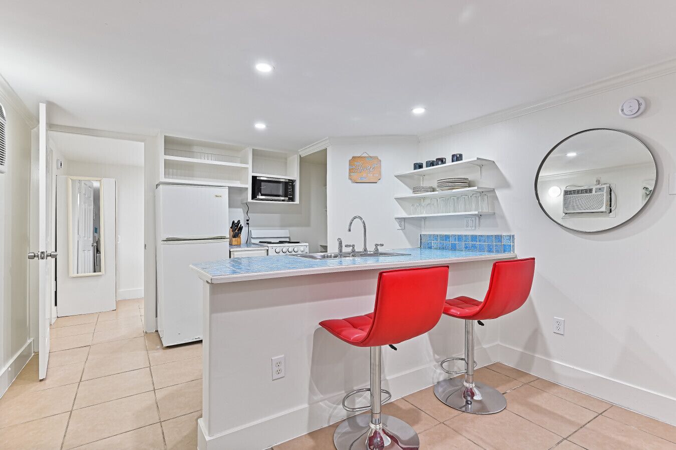 Una cocina compacta con una isla central, microondas, estufa y pileta. Hay dos taburetes rojos y un espejo redondo en la pared.