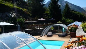 A pool with a glass enclosure, wooden deck, outdoor seating, and a mountain backdrop. - Holiday Home in Champagny Near Vanoise (Champagny-en-Vanoise)