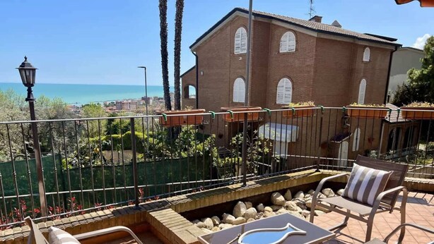 Une terrasse avec du mobilier d’extérieur, une vue sur la mer et, au fond, un bâtiment en briques.