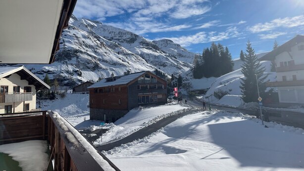 A snow-covered mountain landscape with buildings, including a wooden cabin and a modern house with a ski lift in the background.
