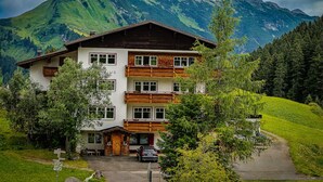 A two-story chalet with a balcony, surrounded by lush greenery and a mountain backdrop.