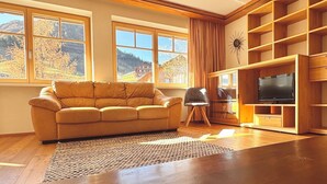 A living room with a sofa, a TV, wooden shelves, and large windows with a view of mountains.