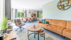 A modern living room with a sofa, armchair, and round wooden table. There is a dining area in the background with a table and chairs. - Luxury 8-person Holiday Villa (De Koog)