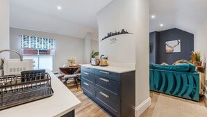 A modern kitchen with a built-in stove, a dining area with a table and chairs, and a teal sofa in a living area. - Glasswing Stays | The Holgate Sanctuary (York)