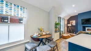 A modern kitchen with a dining area, featuring a wooden table set for four, a blue accent wall, and a large window with blinds. - Glasswing Stays | The Holgate Sanctuary (York)