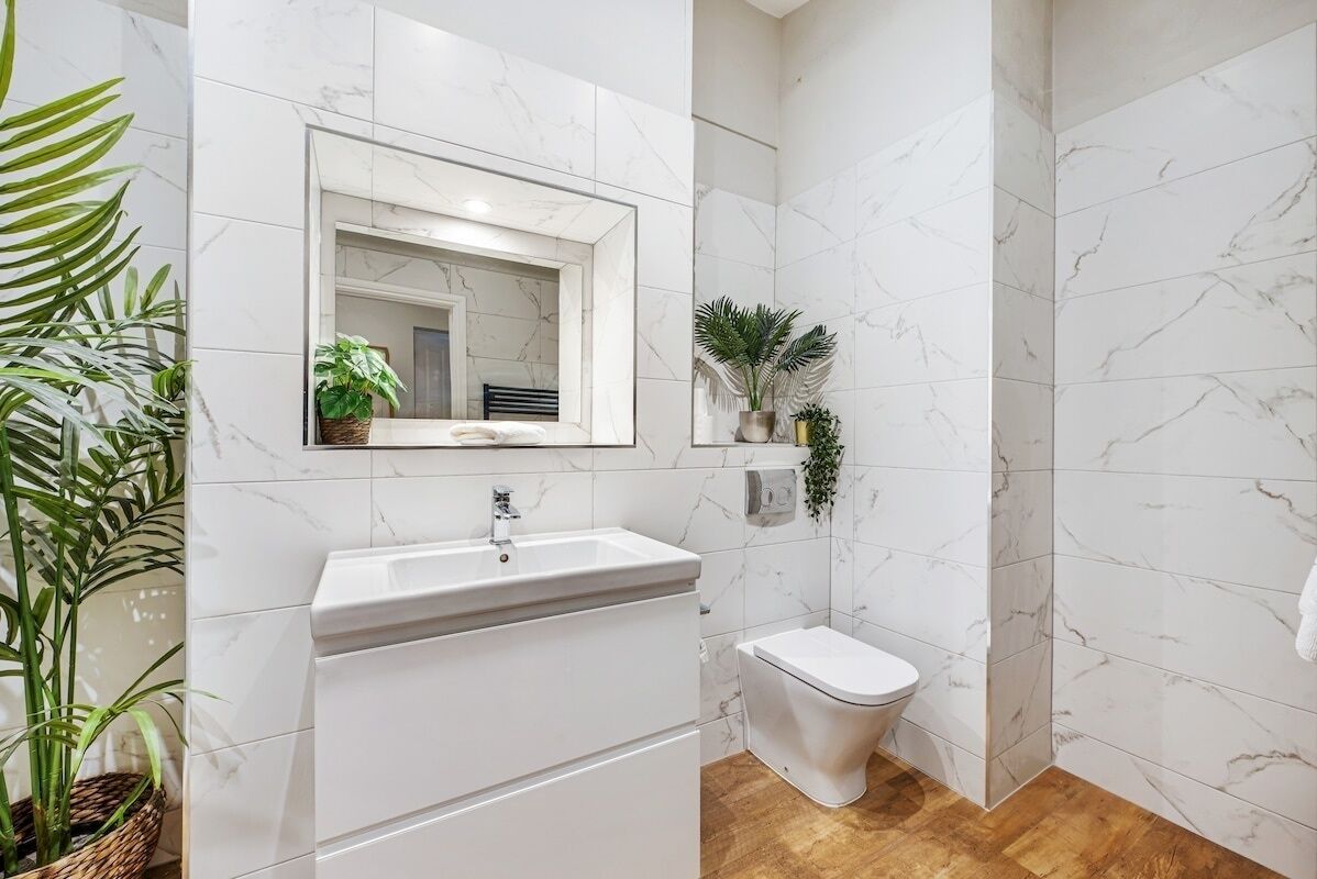 A bathroom with a white sink, a toilet, and a mirror. There are plants on the shelves and a wooden floor.