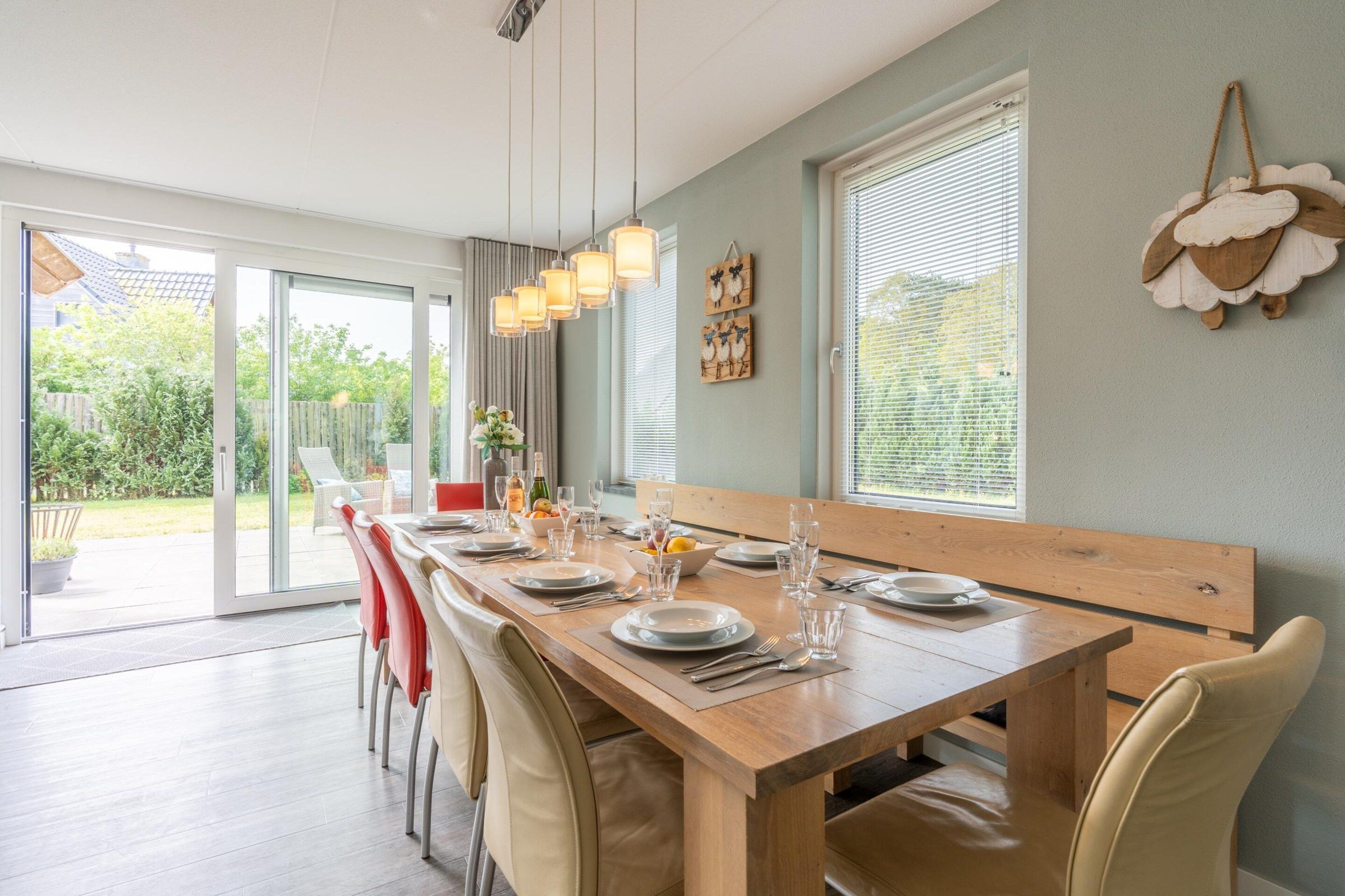 A dining area with a long wooden table set for six, surrounded by chairs, with a view of the garden through large windows.