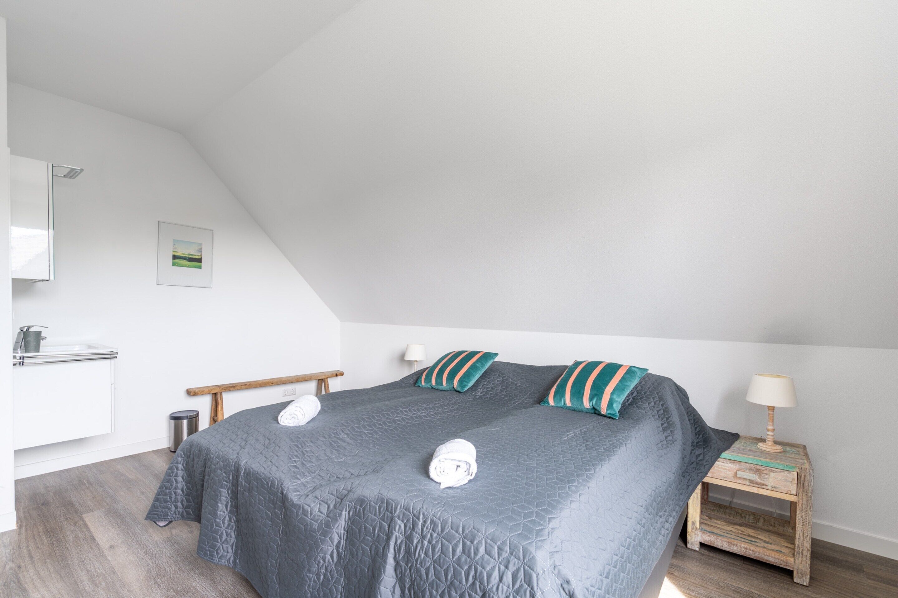 Una camera da letto con un letto grande, un comodino con una lampada, una panca in legno, un cestino e un armadietto bianco con uno specchio.