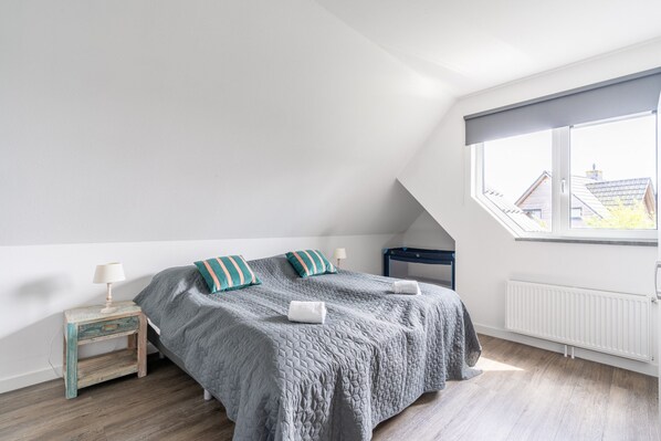 Un dormitorio con techo inclinado, una cama con colcha gris, dos almohadas, una mesita de noche con lámpara y una ventana con vistas a edificios.