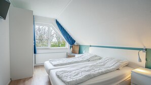 Quarto com duas camas, janela com cortinas azuis, mesa de cabeceira com abajur e radiador.