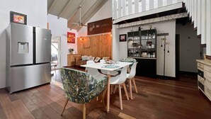Une cuisine spacieuse dotée d’un réfrigérateur moderne en acier inoxydable, d’un coin repas avec une table et des chaises blanches, et d’un plafond en bois avec un escalier apparent.
