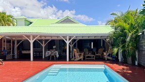 Une piscine avec une terrasse en bois, un patio couvert doté de chaises et de tables, et un bâtiment surmonté d’un toit végétalisé.