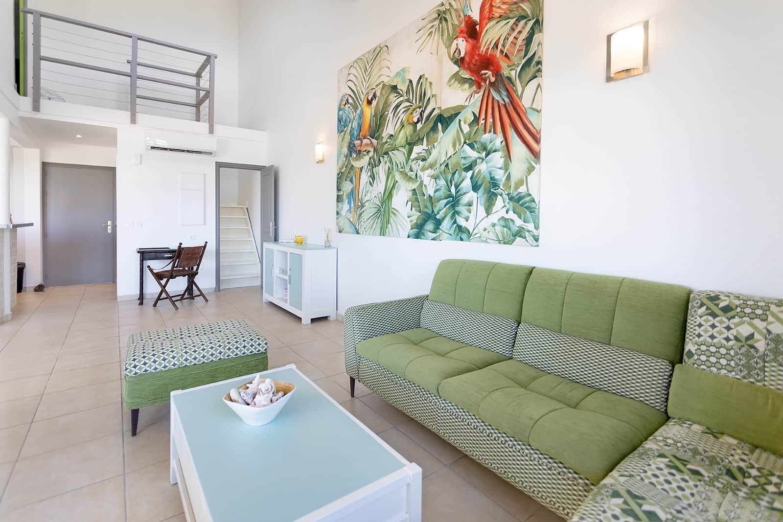 A spacious living area with a green sofa, a patterned ottoman, a dining table with chairs, and a large tropical mural on the wall.