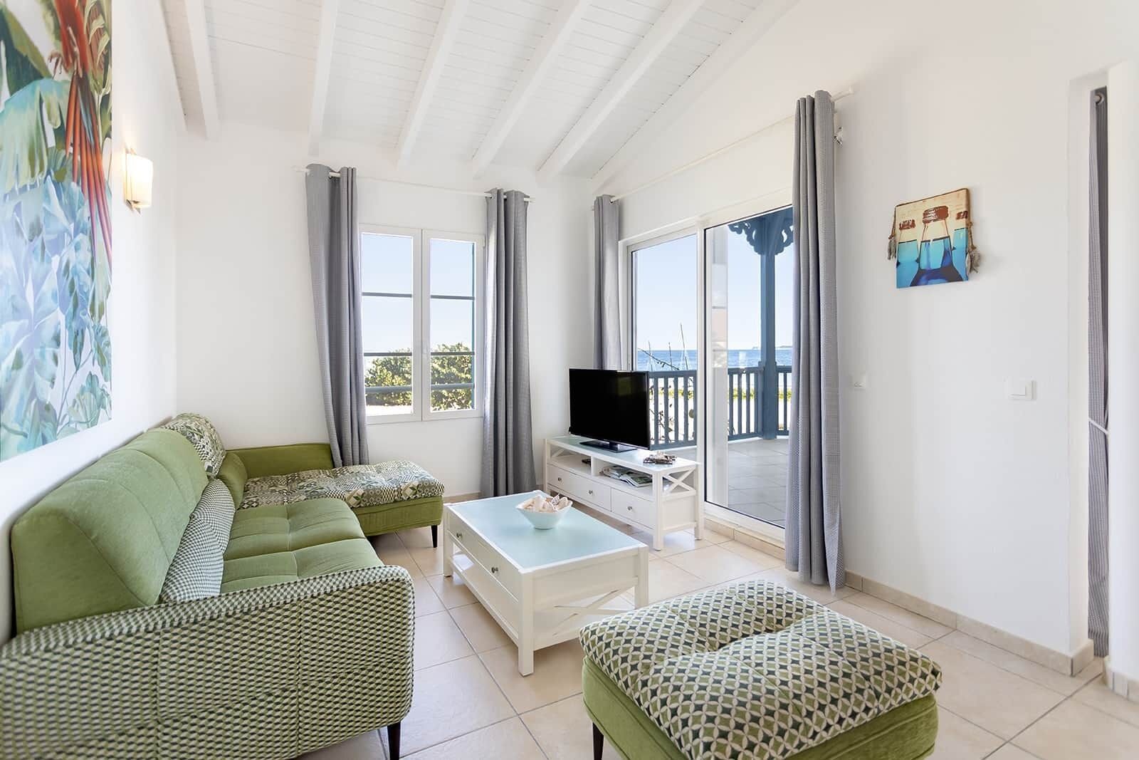 A spacious living room with a green sofa set, a coffee table, and a TV on a white stand. Large windows offer a view of the sea.
