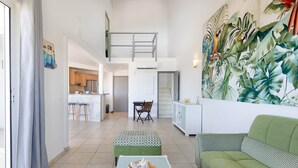 A spacious living area with a green sofa, a patterned ottoman, and a dining table. A kitchen is visible in the background, and a staircase leads to an upper level. - Timoon, 2 bedroom apartment on the beach with amazing view! (Marigot)