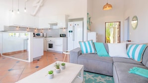 A modern kitchen with white cabinets, a dining area with a grey sofa and striped cushions, and a small table with potted plants.
