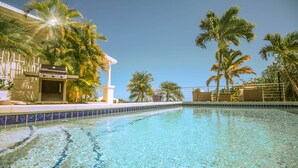 A swimming pool with clear water, surrounded by palm trees and a building with a barbecue area.