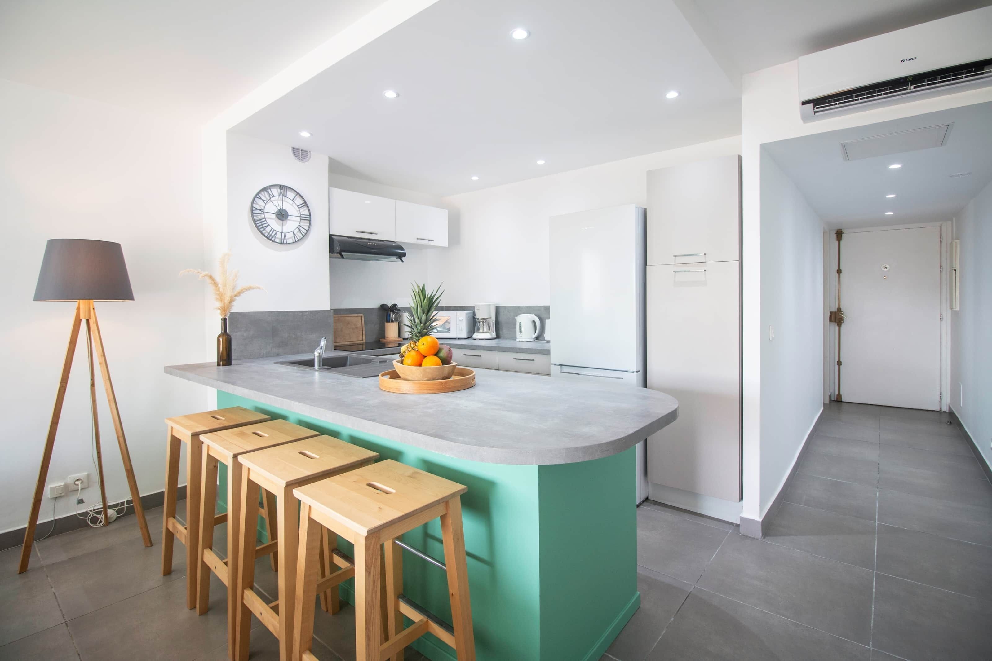 A modern kitchen with a central island, wooden stools, a clock, and a large window.