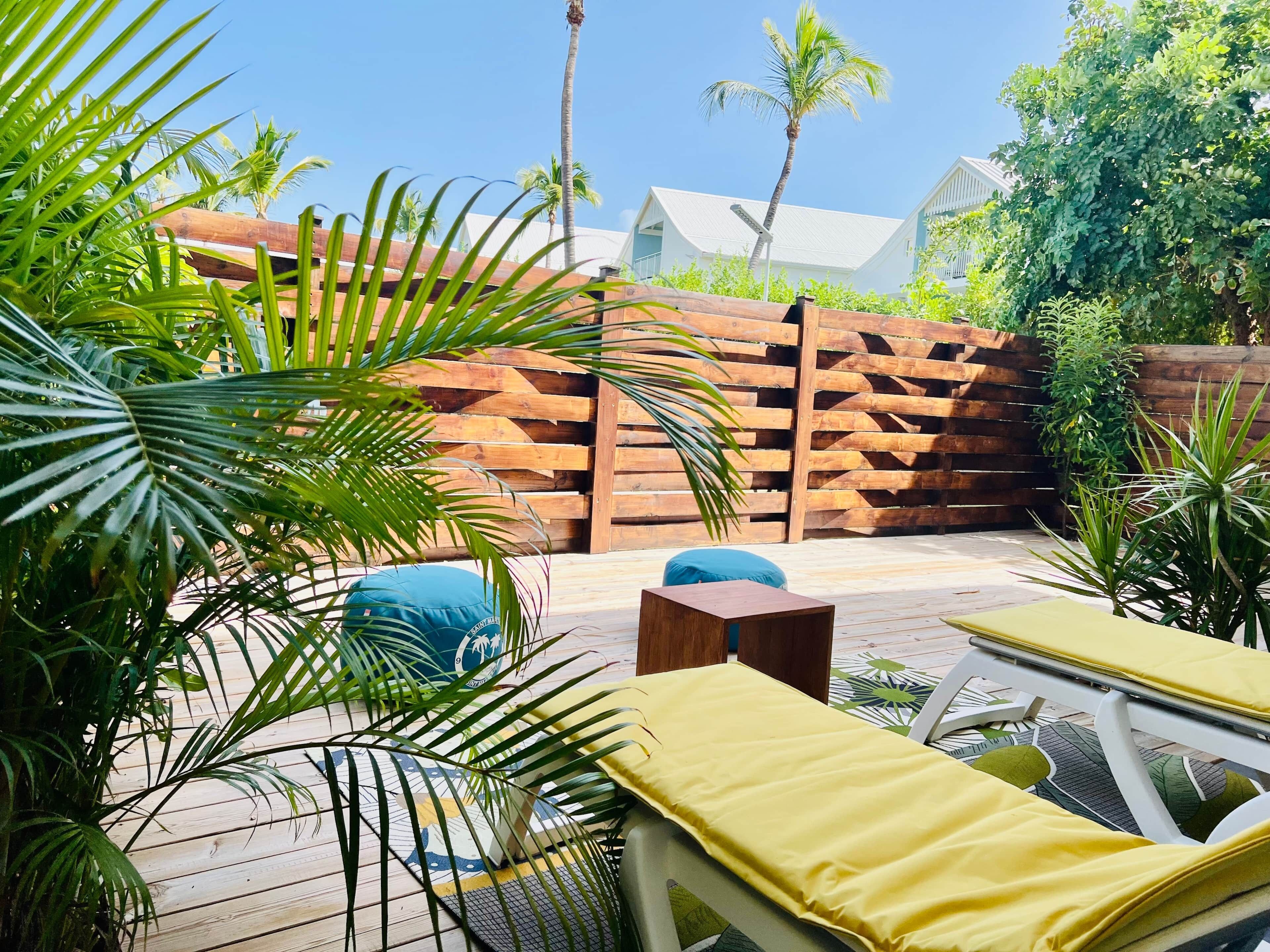 A patio area with wooden furniture, including a lounge chair and a small table, surrounded by lush greenery and a wooden fence.