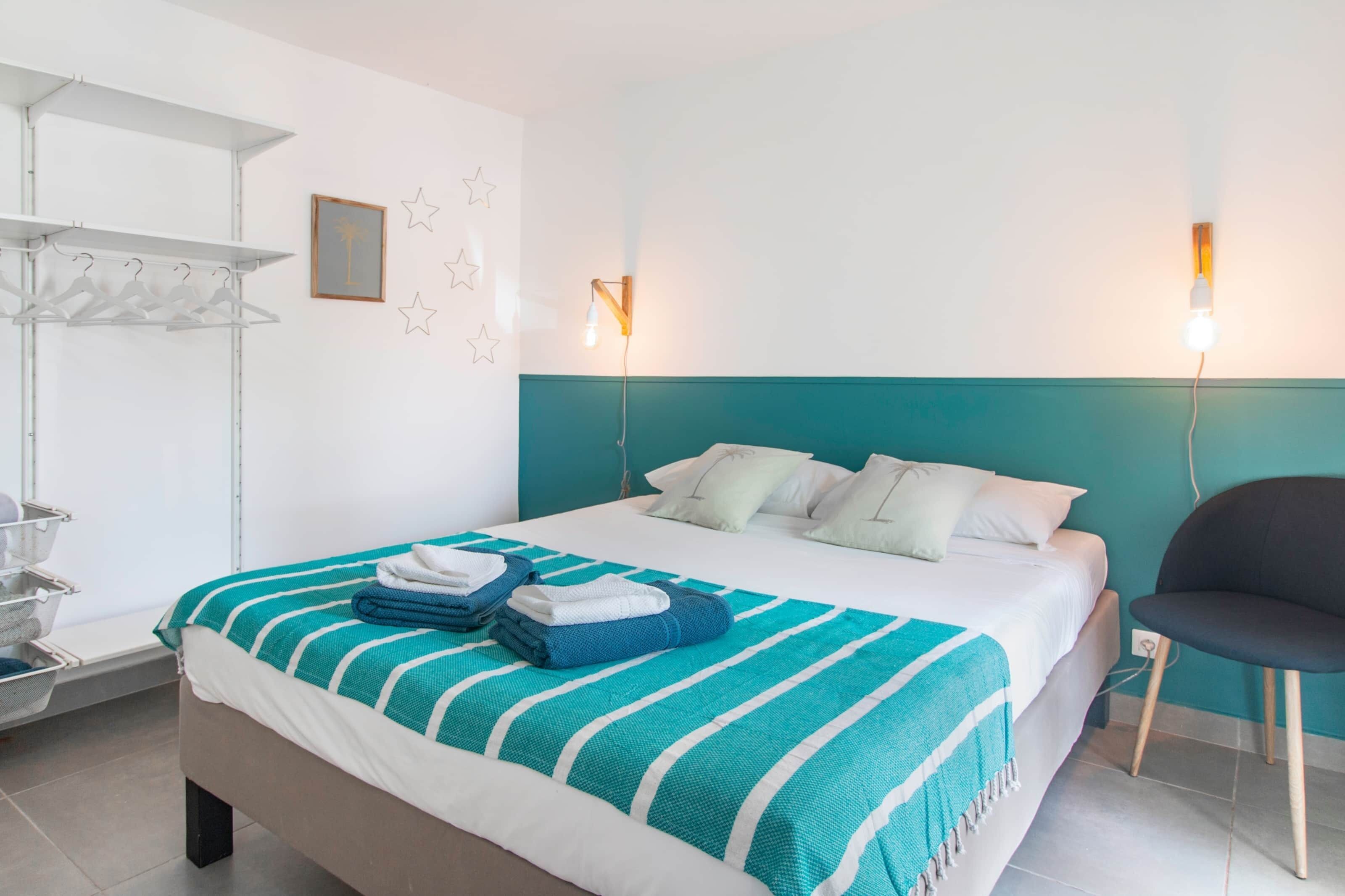 Una cama bien hecha con una manta a rayas en tonos aguamarina y blanco, una silla y una lámpara fijada a la pared.