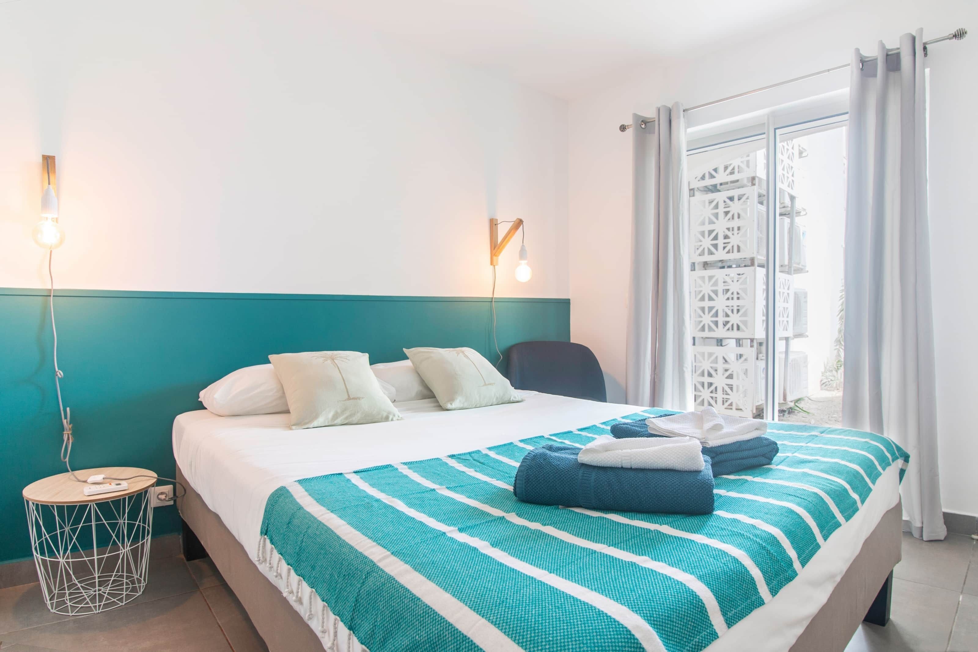 Habitación de hotel con cama, una mesita redonda, luz de pared y vista al exterior a través de una ventana con cortinas.