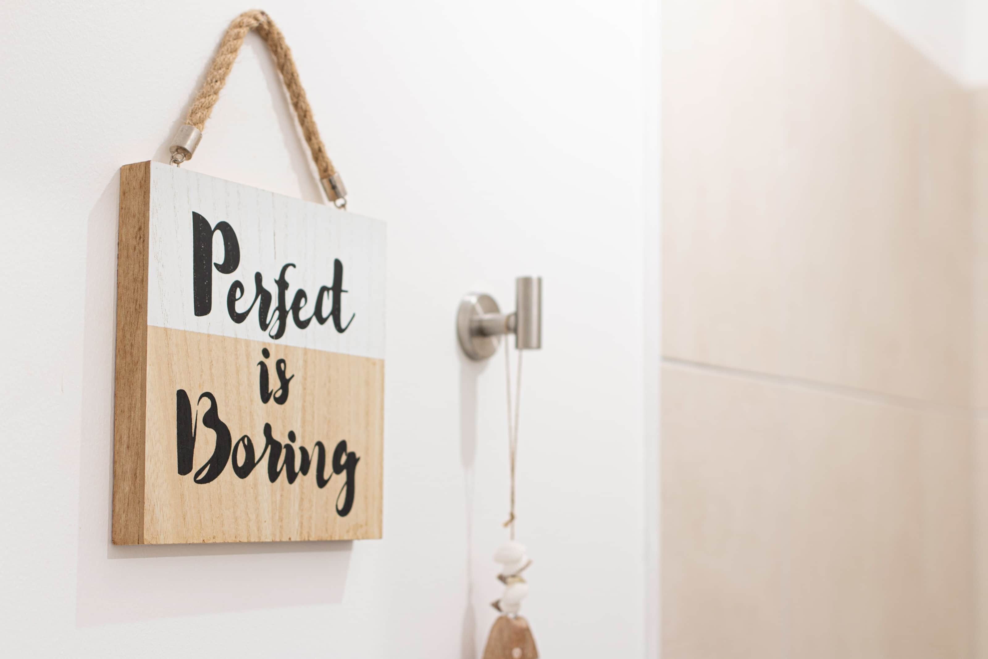 A wooden sign with the text "Perfect is Boring" hanging on a white door.