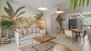 A modern living room with a sofa, coffee table, and dining area. There is a staircase leading to another level, a kitchen, and a large mural on the wall. - Maison GAMA, 2 bedrooms and shared pool (CISTERN!!!) (Collectivity of Saint Martin)