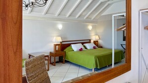A bedroom with a bed, bedside lamps, a wooden headboard, a wicker chair, and a ceiling with wooden beams. - Studio Palm Court with shared pool behind Orient Beach (Collectivity of Saint Martin)