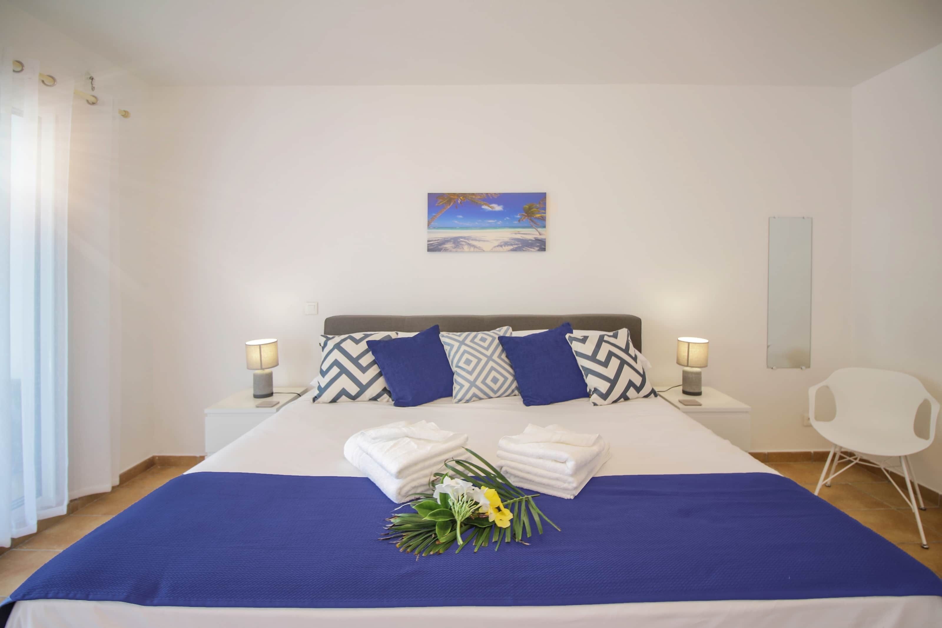 Un letto rifatto con una coperta blu, due cuscini e un bouquet di fiori. Sono presenti due comodini con lampade e una sedia bianca.