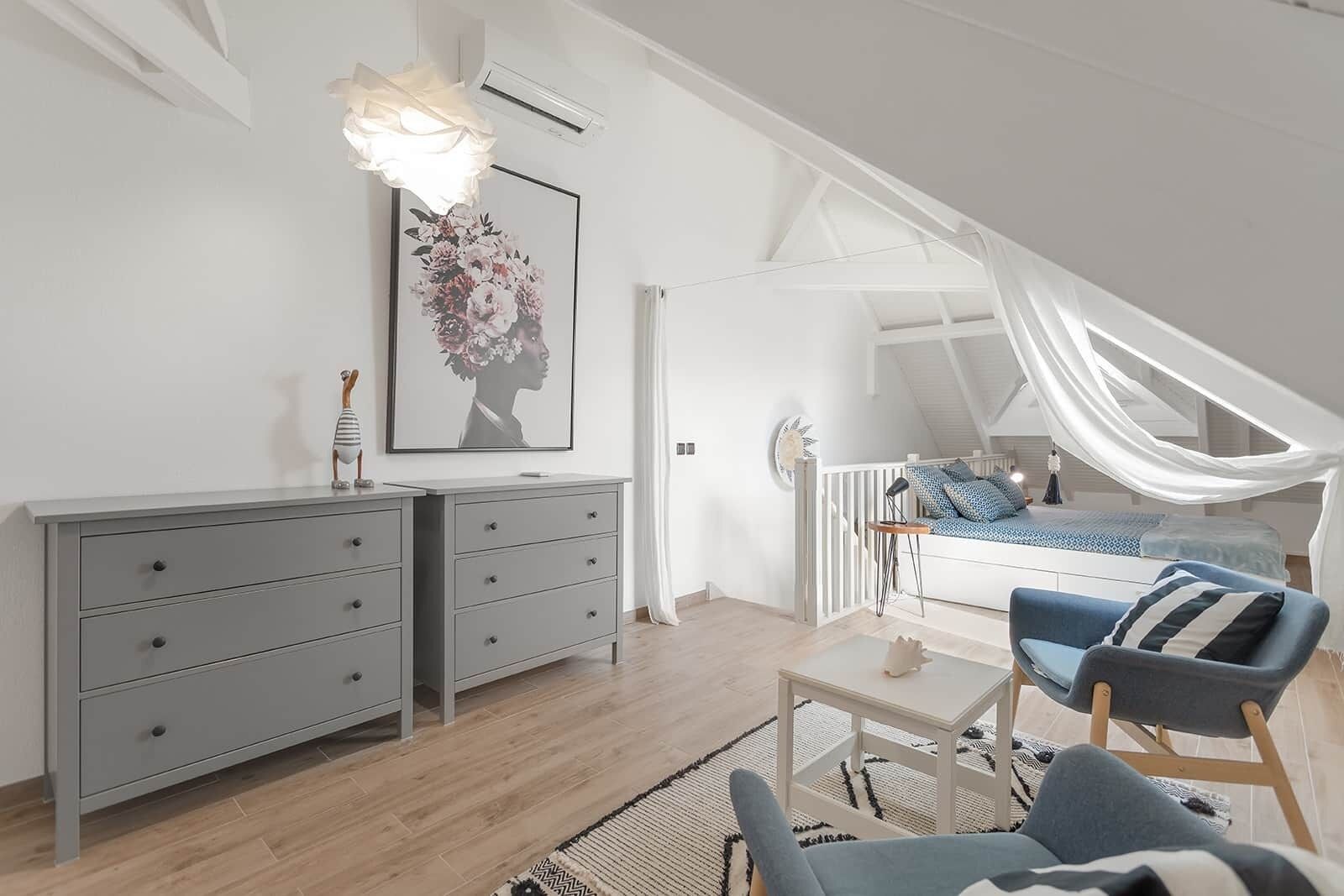 Ett modernt sovrum i loftstil med ett sluttande tak, en säng med himmelssäng, en grå byrå och en blå fåtölj.